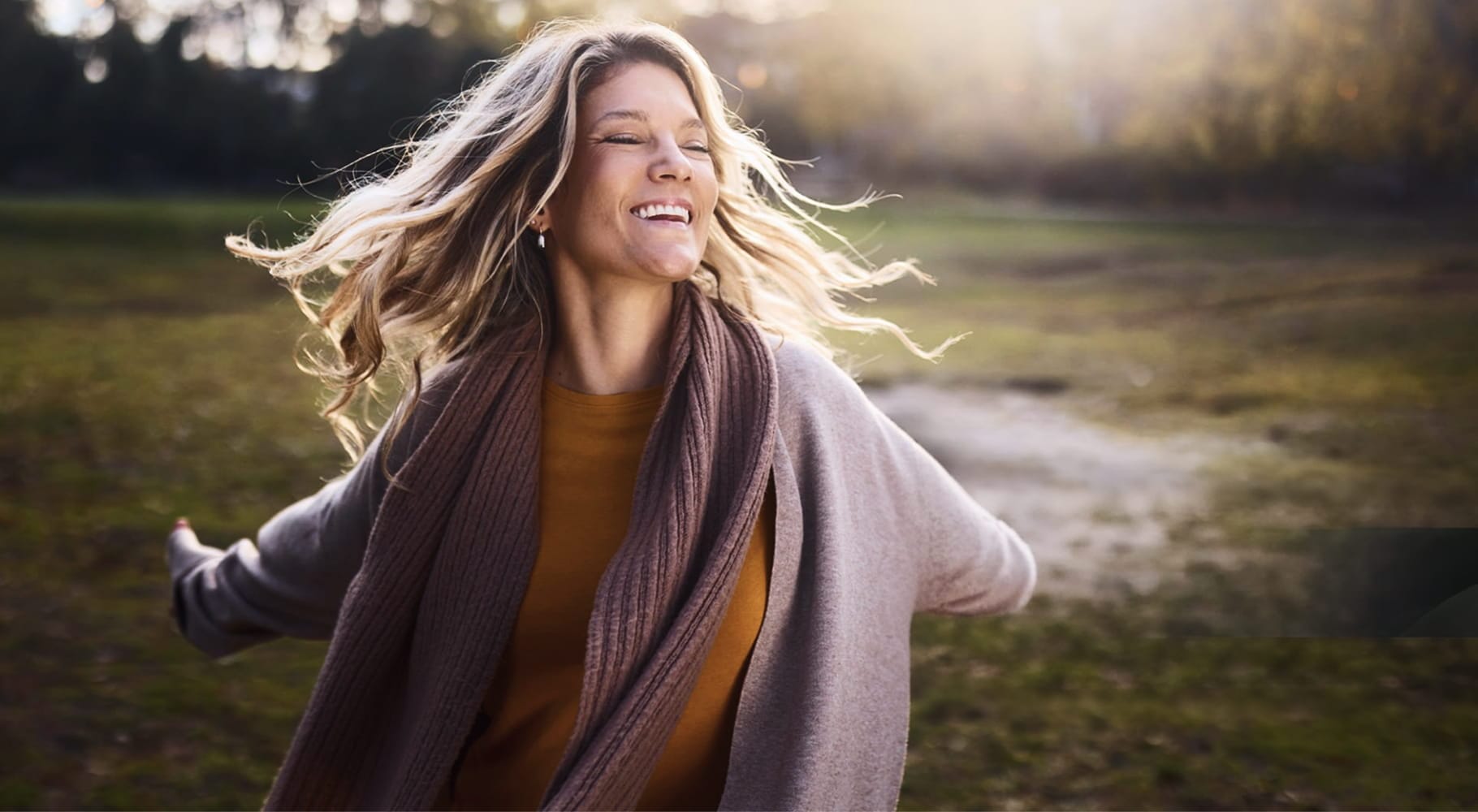 a cheerful woman wearing a brown ribbed scarf and a gray draped cardigan over a mustard yellow top, capturing a moment of bliss in a park
