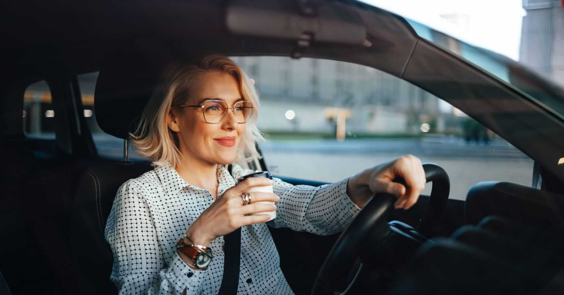 a woman with blond hair wearing glasses driving a car and holding a cup